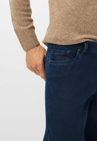 Jean en denim bleu marine avec une coupe ajustée, dotés d'une poche avant et de détails de rivets discrets, associés à un pull en tricot marron clair.