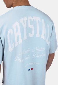 Světle modré tričko s výrazným bílým blokovým textem "CRYSTAL" a kurzívou "Nordic Nights Dive into Darkness." Obsahuje malý detail francouzské vlajky.