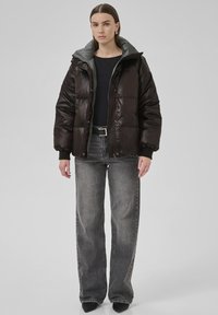 Kvinde iført en mørkebrun puffjakke over en sort top, grå brede jeans og sorte spidse sko foran en ensfarvet baggrund.