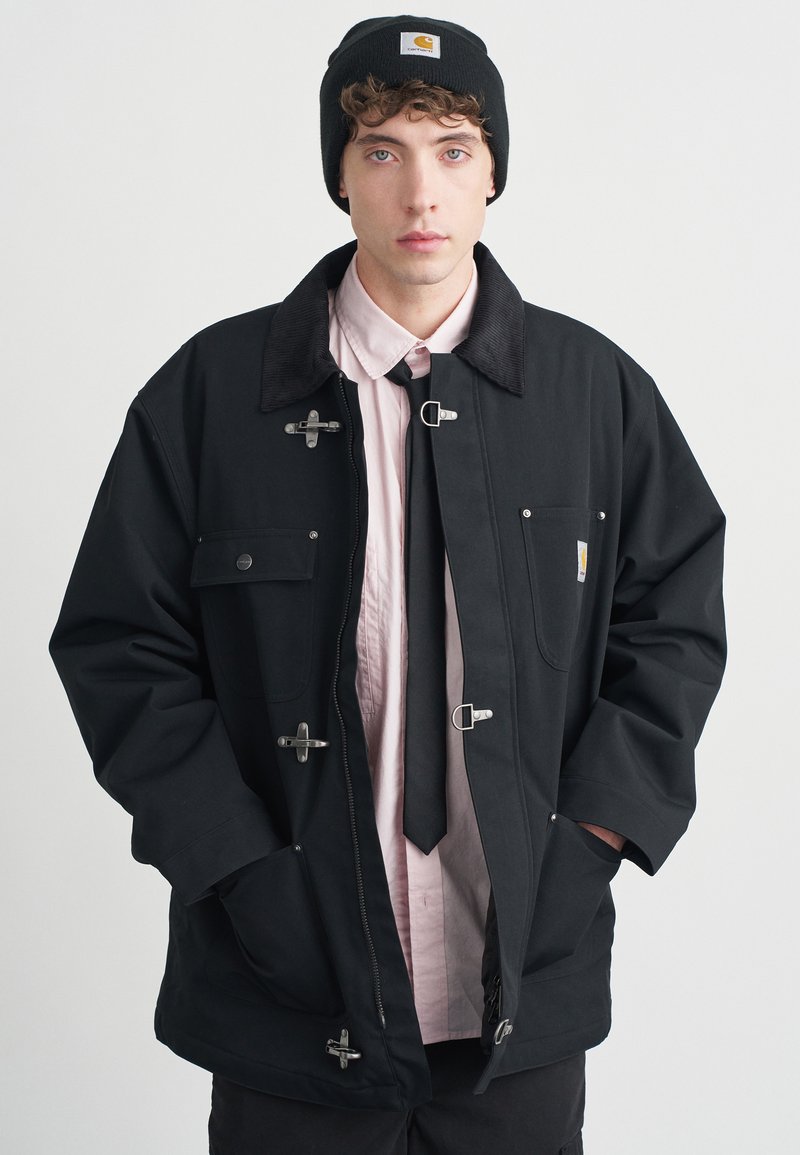 Giacca nera con dettagli in argento, ampio colletto e tasche frontali. Indossata su una camicia rosa chiaro e cravatta nera, abbinata a un berretto nero.