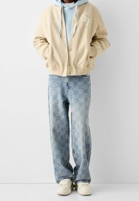 Beżowa kurtka zapinana na napy, pod nią jasnoniebieska bluza z kapturem, wzorzyste jasnoniebieskie dżinsy i białe sneakersy. Miękki materiał, luźny krój i wzór w kratkę.