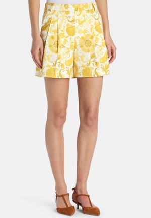 Kvinde iført hvide shorts med gult blomstermønster og brune T-rem hæle, stående mod en ensfarvet baggrund.