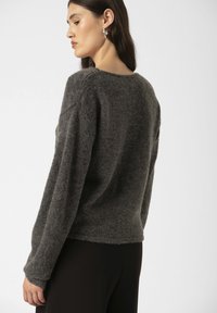 Modelul este văzut din spate, purtând un pulover texturat de culoare gri închis și pantaloni negri, cu părul lung lăsat liber și cercei rotunzi din argint.