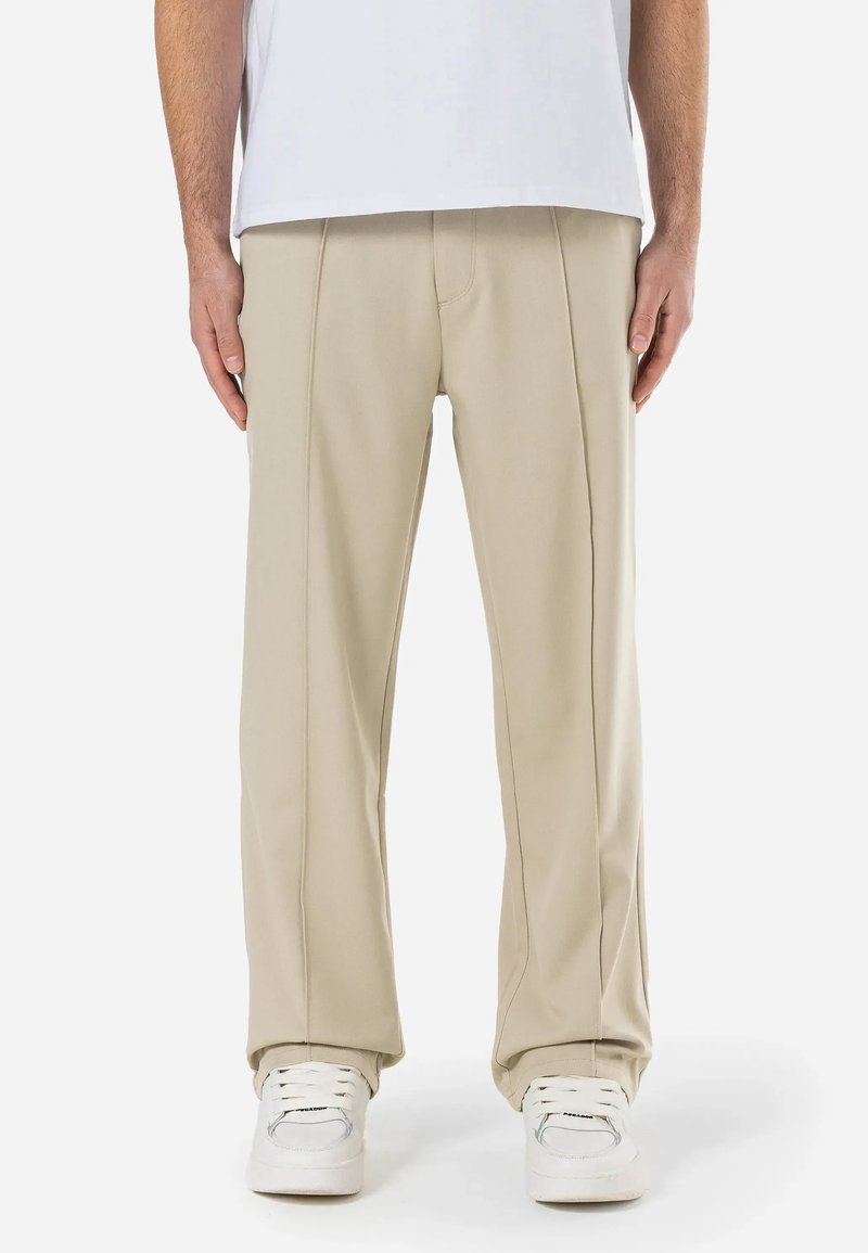 Homme portant un pantalon beige à jambes droites et des baskets blanches, debout avec les bras détendus le long du corps devant un fond uni.