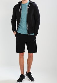 Sudadera con cremallera negra, camiseta de manga corta color azul verdoso, shorts negros y zapatillas negras. Presenta cordones y acentos de logo en contraste.