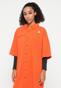Camicia arancione con colletto, maniche corte e tasca frontale. Presenta un piccolo logo e una vestibilità comoda. Tessuto leggermente lucido.