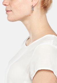 Orecchino a cerchio argentato mostrato su un modello con pelle chiara. L'orecchino ha una superficie liscia e riflettente e un design minimalista. Il modello indossa una camicia bianca.