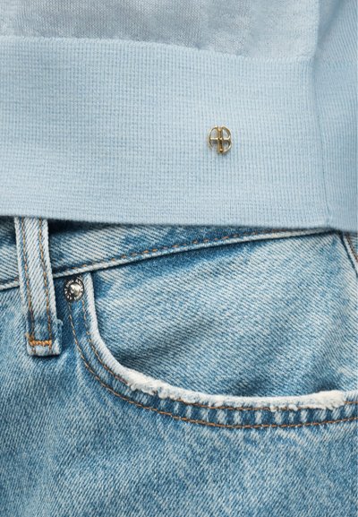 Lichtblauwe gebreide trui met klein gouden logo boven de tailleband, gecombineerd met lichtblauwe spijkerbroek met zichtbare voorzak.