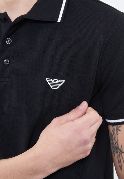 Man draagt een zwart poloshirt met witte gestreepte kraag en manchetten, met een wit geborduurd logo op de borst; tatoeage zichtbaar op de onderarm.