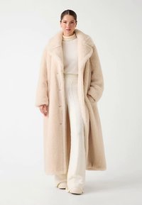 Manteau en fausse fourrure beige avec un large col, des boutons sur le devant, des manches longues et un design fluide, associé à une tenue tricotée crème.