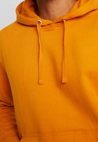 Sudadera con capucha de color naranja brillante, con cordones y un bolsillo tipo canguro, usada por una persona, enfocándose en el área del pecho y el cuello.