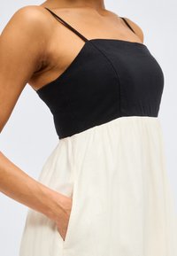 Γυναίκα που φοράει ασπρόμαυρο φόρεμα χωρίς μανίκια με λεπτές τιράντες και το χέρι της τοποθετημένο στην πλευρική τσέπη του φορέματος.