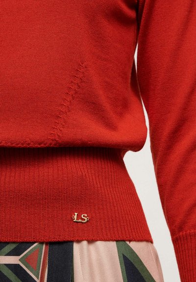 Raudonas megztas džemperis su raukšlėta apatine dalimi ir dekoratyvine siuvinėjimu. Prie apatinės dalies yra nedidelis auksinis "LS" logotipas. Lygus paviršius.