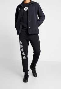 Hombre vestido con chaqueta negra de Adidas, pantalones negros con diseños de logo blanco a lo largo de la pierna, zapatillas negras, de pie contra un fondo claro y sencillo.