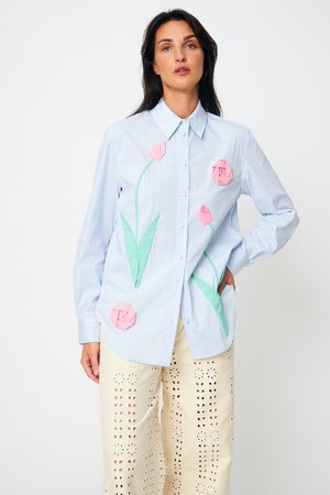 Naine, kes kannab helehalli triibulist särkiga, millel on 3D roosad lilled ja rohelised lehed, mida on kombineeritud kreemikõrgete laia säärega pükstega, millel on perforatsioon.