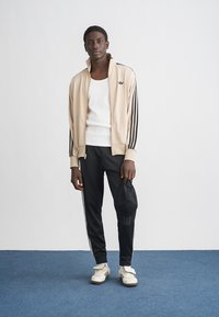 Bež jakna na patent s crnim prugama, bijeli top, crne sportske hlače, bijele tenisice i crna kapa, stoji na plavom podu.