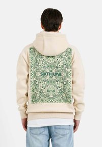 Sudadera beige con un diseño de paisley verde en la parte trasera. Presenta capucha con cordones, puños acanalados y un logo, fabricada con un tejido suave.