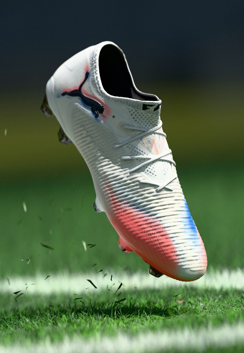 White soccer cleat with a textured upper, featuring pink and blue accents, and a Puma logo. Positioned above grass, capturing motion.