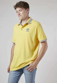 Polo shirt jaune avec un col bleu et à motifs, arborant un logo sur la poitrine et des manches courtes. Tissu doux et design décontracté.