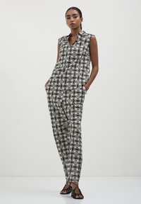 Donna che indossa un top senza maniche geometrico bianco e nero e pantaloni abbinati, con tacchi neri a fascia e bracciale argentato, su uno sfondo bianco uniforme.