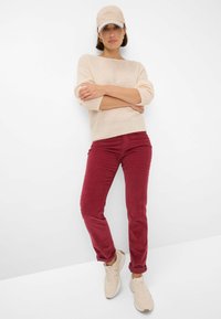 Un pull en tricot beige, un pantalon en velours côtelé bordeaux et des baskets claires. L'accessoire comprend une casquette beige. Design simple avec des textures douces.