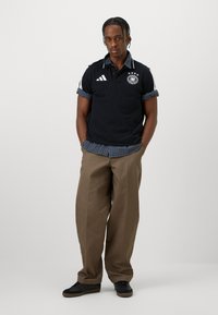 Mladý muž s pletenými vlasy stojí s rukama v kapsách, obléká černé polo tričko, modrou kostkovanou košili, hnědé kalhoty a černé tenisky na bílém pozadí.