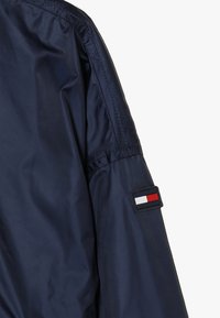 Giacca blu navy con texture lucida. Presenta una patch con il marchio sulla manica in rosso, bianco e blu. Dettagli delle cuciture lungo il braccio.