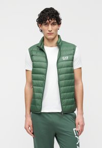 Grön vadderad väst med hög krage, lätt quiltad design och varumärkeslogotyp på bröstet, lager-på-lager över en vit t-shirt.