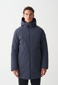 Muž v tmavomodrom zimnom kabáte s kapucňou a predným zipsom, stojaci pred hladkým bielym pozadím.