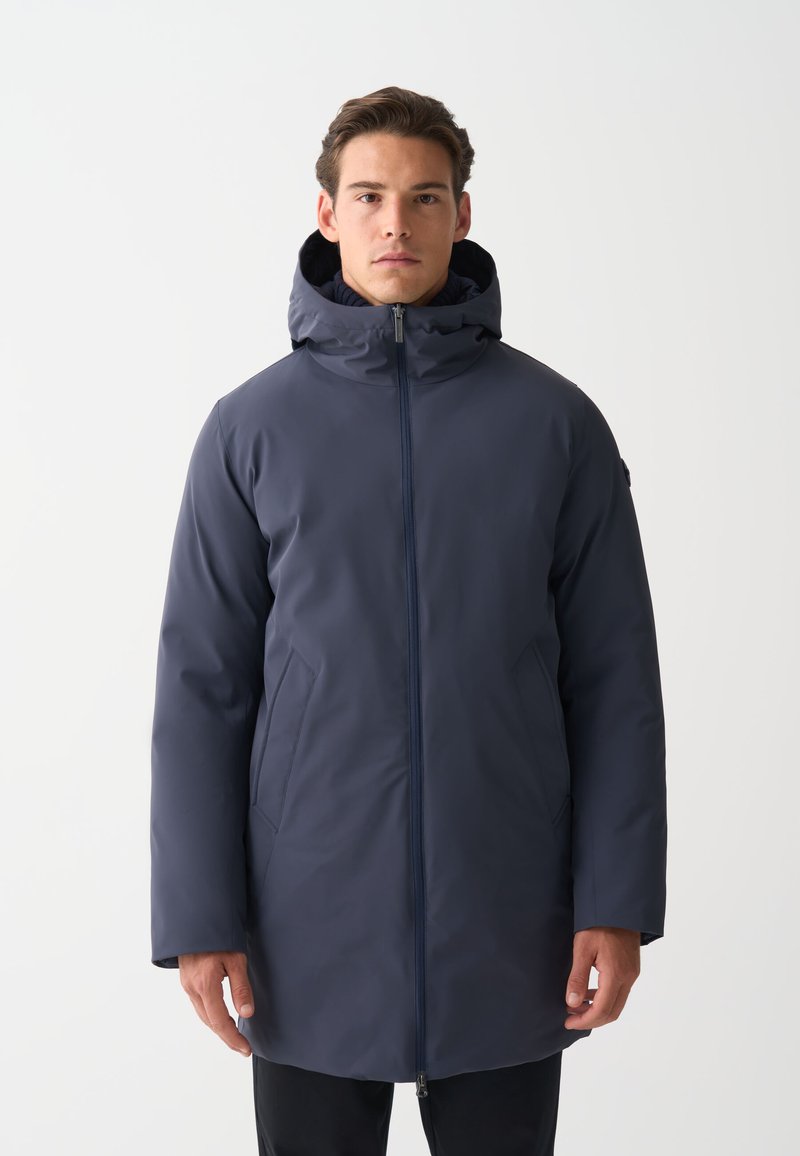 Muž v tmavomodrom zimnom kabáte s kapucňou a predným zipsom, stojaci pred hladkým bielym pozadím.