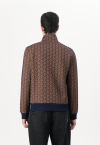 Brun mønstret jakke med høj krave, navy accenter, ribbede manchetter og kant. Glat tekstur med geometriske designs gennem hele jakken.
