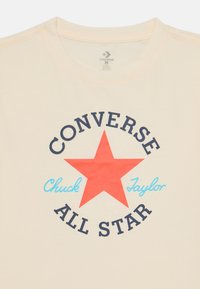 Camiseta de algodón color crema con una estrella roja y el texto "CONVERSE ALL STAR" en azul oscuro y azul claro. Diseño clásico de cuello redondo.
