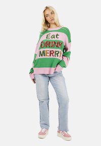 Maglione a righe verdi e rosa con testo in paillettes "Eat DRINK MERRY!" in rosso, oro e verde, abbinato a jeans azzurri e sneakers rosa.