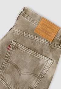 Μπεζ παντελόνι τζιν με δερμάτινο ένθετο με την ετικέτα «Levi Strauss & Co.», λεπτομερείς ραφές στις τσέπες και κόκκινη υφασμάτινη ετικέτα στο πίσω μέρος.