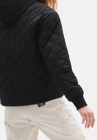 Czarna pikowana kurtka z kapturem, z teksturowanym wzorem i ściągaczami na mankietach, noszona na kremowych spodniach typu cargo z widocznymi kieszeniami i oznaczeniami marki.