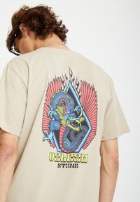 Beige cotton t-shirt featuring a colorful dragon graphic with red wings and blue accents, positioned inside a diamond shape.