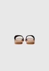 Svart vevde slidesandaler med flat tan såle, med en glatt tekstur og minimalistisk design, sett fra baksiden.