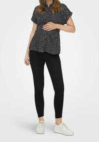 Blouse noire à manches courtes avec un motif et un col à boutons, associée à un jean skinny noir et des baskets blanches.