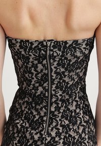 Utan axelklänning med svart blommönstrad spetsöverdrag och synlig silverblixtlåsstängning i ryggen på bar hud.