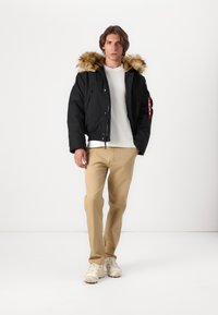 Black bomber jacket with a fur-lined hood, zip front, and side pockets; paired with a white shirt, beige pants, and light shoes.