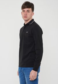 Polo shirt nero a maniche lunghe con profilo bianco del colletto, caratterizzato da un logo sul petto e una fila di bottoni, abbinato a jeans denim azzurri.