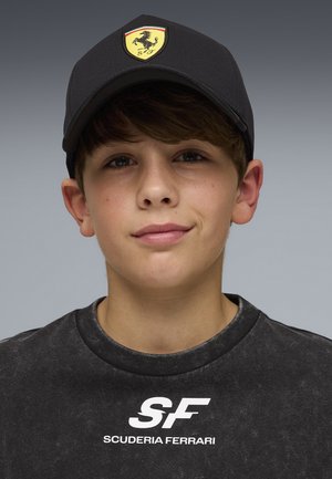 Teenager-Junge trägt eine schwarze Scuderia Ferrari Kappe und ein passendes schwarzes Sweatshirt mit weißem SF-Logo vor einem schlichten grauen Hintergrund.