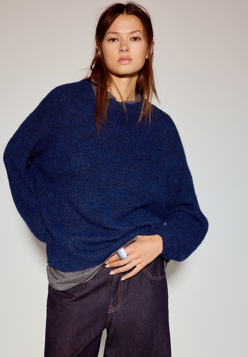 Oversized blauwe gebreide trui met een gestructureerde stof, verlaagde schouders en lange mouwen, gecombineerd met een grijze onderlaag en donkere jeans.