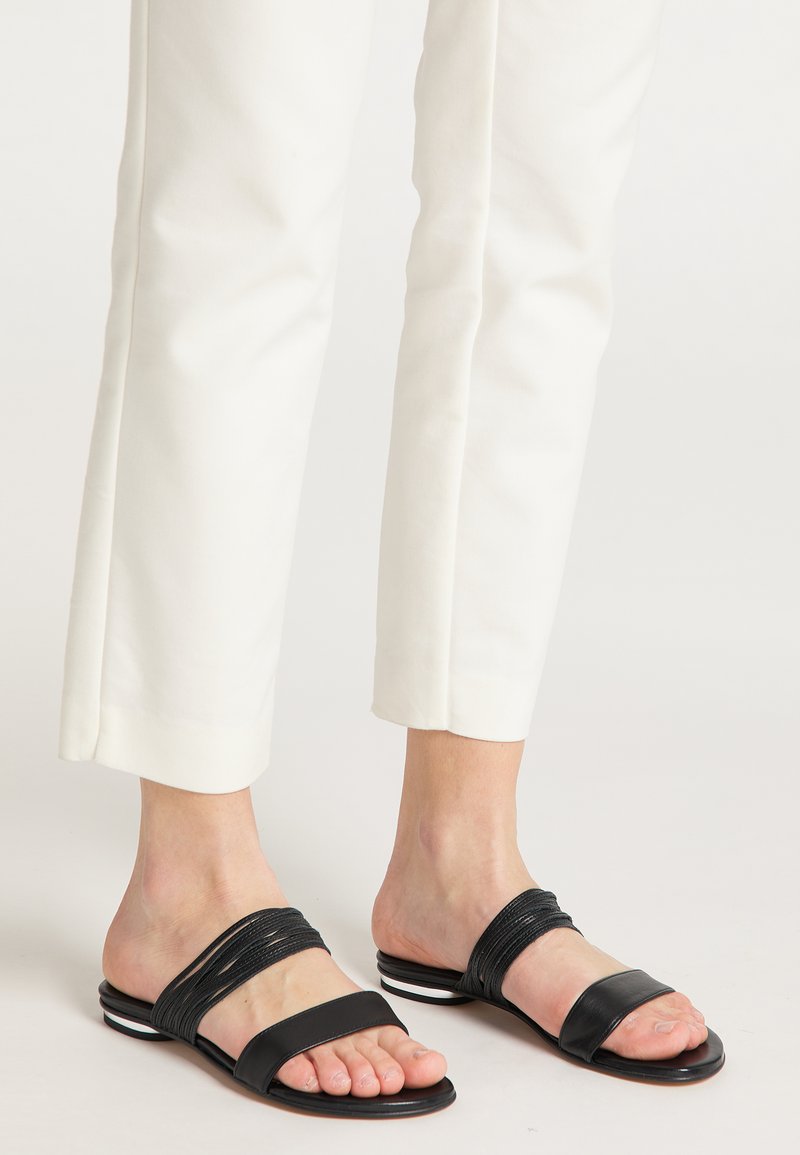 Zwarte slip-on sandalen met meerdere gestructureerde banden, open teen ontwerp en een vlakke zool met een subtiele witte accent. Draagt met witte broek.