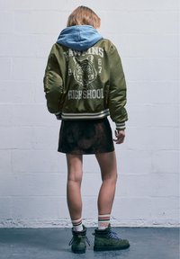 Oliivinvihreä pommikenttätakki, jossa on tiikeri- grafiikka, kerrostettuna vaaleansinisen hupparin päälle. Musta mokkanahkainen mini-hame ja vihreät nauhakengät, joissa on siniset nauhat.