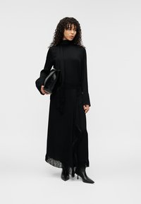 Robe longue noire avec un corsage ajusté, des manches fluides et des détails en franges le long de l'ourlet. Associée à une pochette noire et des bottes à talons.