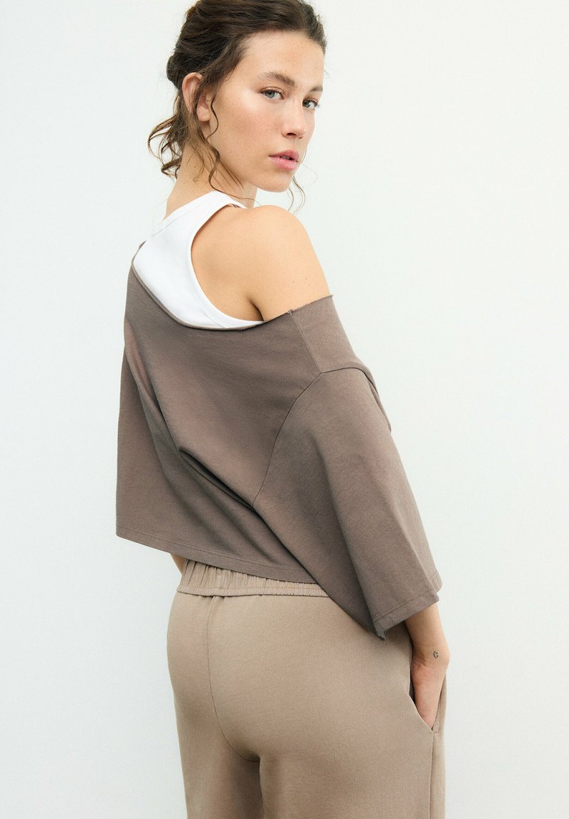 Kvinde med bølget hår i en løs brun off-shoulder top over en hvid tanktop, iført beige bukser, der kigger over sin skulder mod en ensfarvet baggrund.
