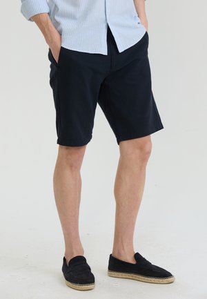 Férfi tengerészkék rövidnadrágban, világoskék csíkos ingben és fekete bebújós cipőben áll, kezét a zsebében tartva.