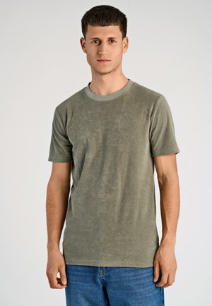 Młody mężczyzna z krótkimi brązowymi włosami, ubrany w oliwkową zieloną koszulę z krótkim rękawem i niebieskie dżinsy, na jasnym jednolitym tle.