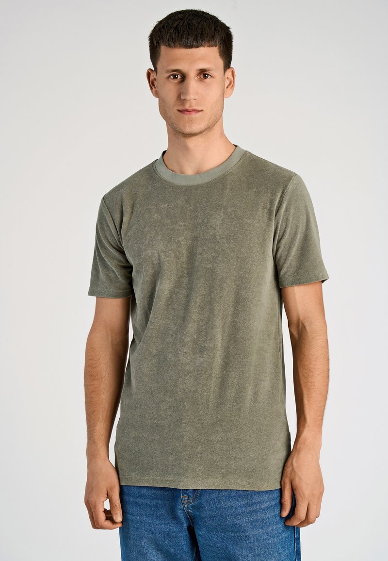 Giovane uomo con capelli corti castani, che indossa una camicia verde oliva a maniche corte e jeans blu, su uno sfondo chiaro e semplice.